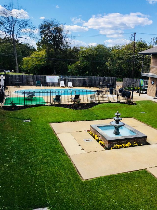 Sunny courtyard lawn with a tiered fountain in a brick basin, with the fenced swimming pool behind