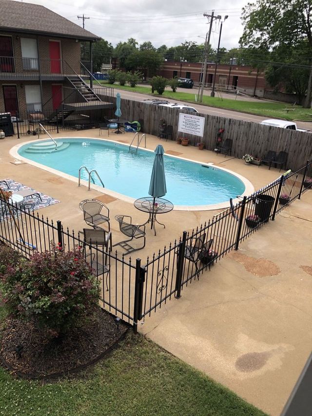 Oval swimming pool with patio chairs, umbrellas, and a black iron fence beside the two-story brick building